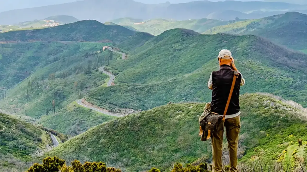 Mann mit Kamera genießt die Aussicht auf kurvige Straße in grünen Bergen. Perfektes Ziel für Wandern in Spanien.