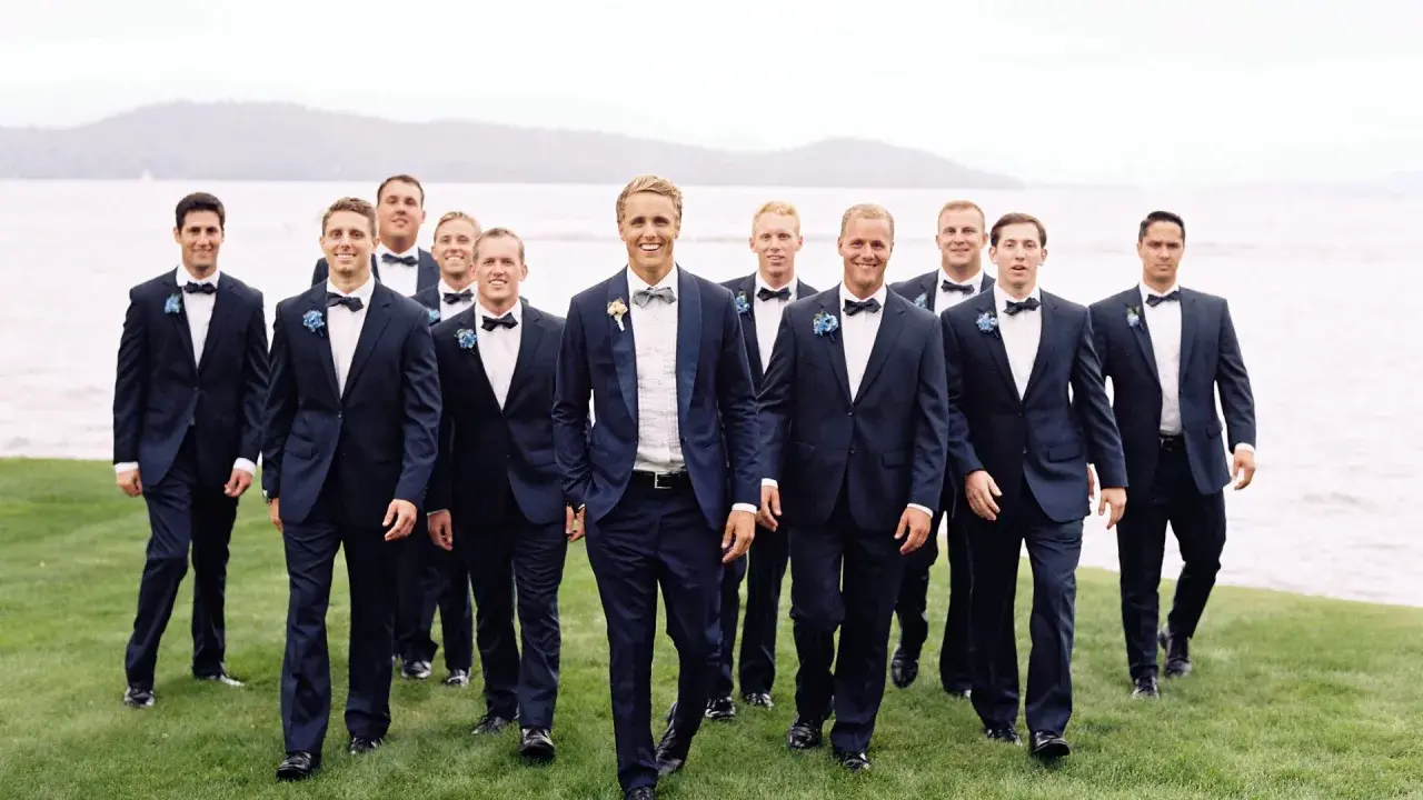 Groomsmen in navy suits with bow ties walk on grass by a lake. Can you wear a normal suit to a black tie event? This image shows a stylish alternative.