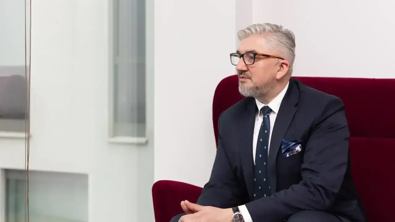 Mężczyzna w garniturze i okularach siedzi na czerwonym fotelu.