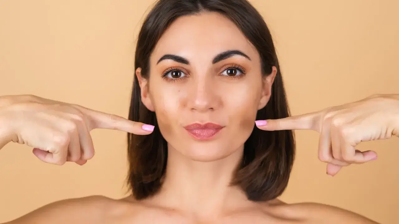 Doigts pointés vers les joues, une femme montre comment repulper son visage pour un look plus jeune et plein.