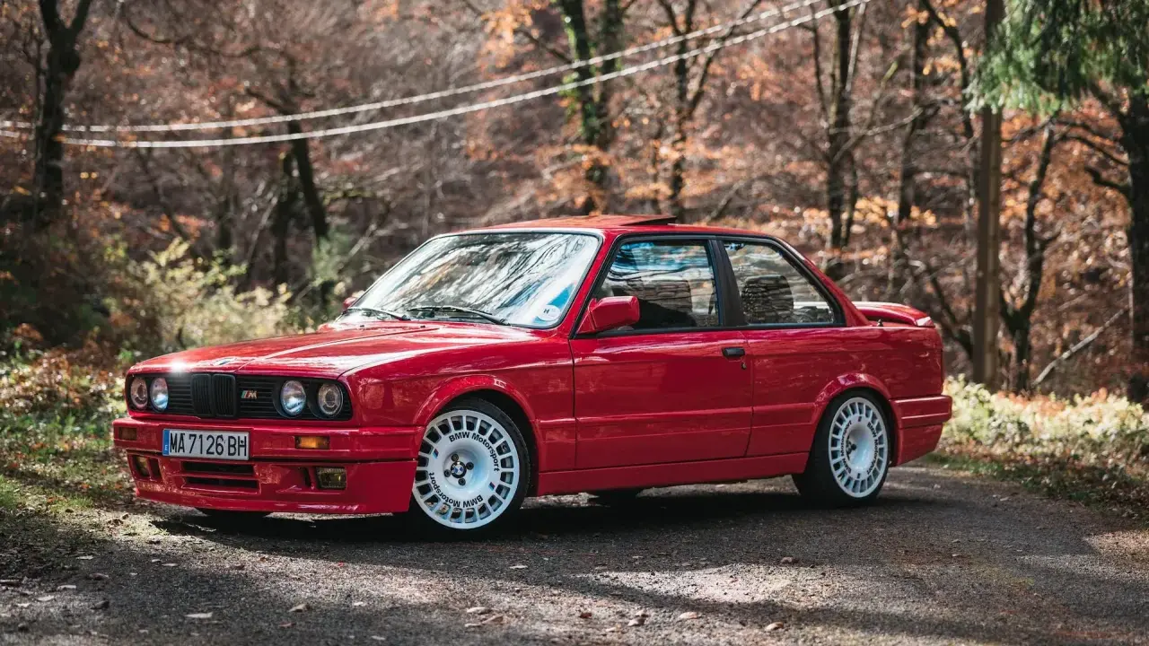 Czerwone bmw e30 coupe z białymi felgami na tle jesiennego lasu.
