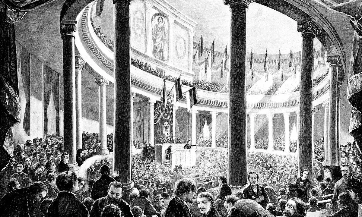 Versammlung der Frankfurter Nationalversammlung in der Paulskirche. Viele Männer in Anzügen und Hemden sitzen und stehen in einem großen, säulenreichen Gebäude.