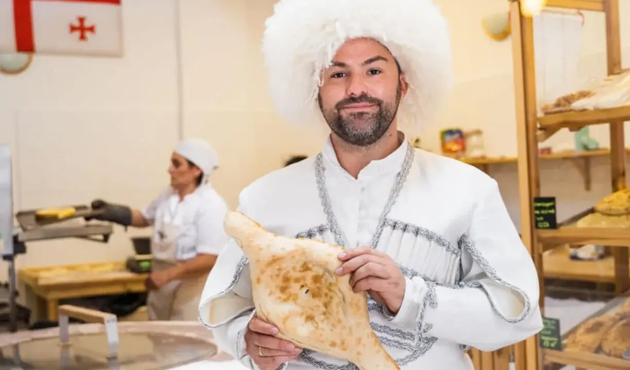 Mężczyzna w tradycyjnym stroju i czapce z owczej wełny trzyma chleb gruziński. W tle widać flagę Gruzji i piekarnię.