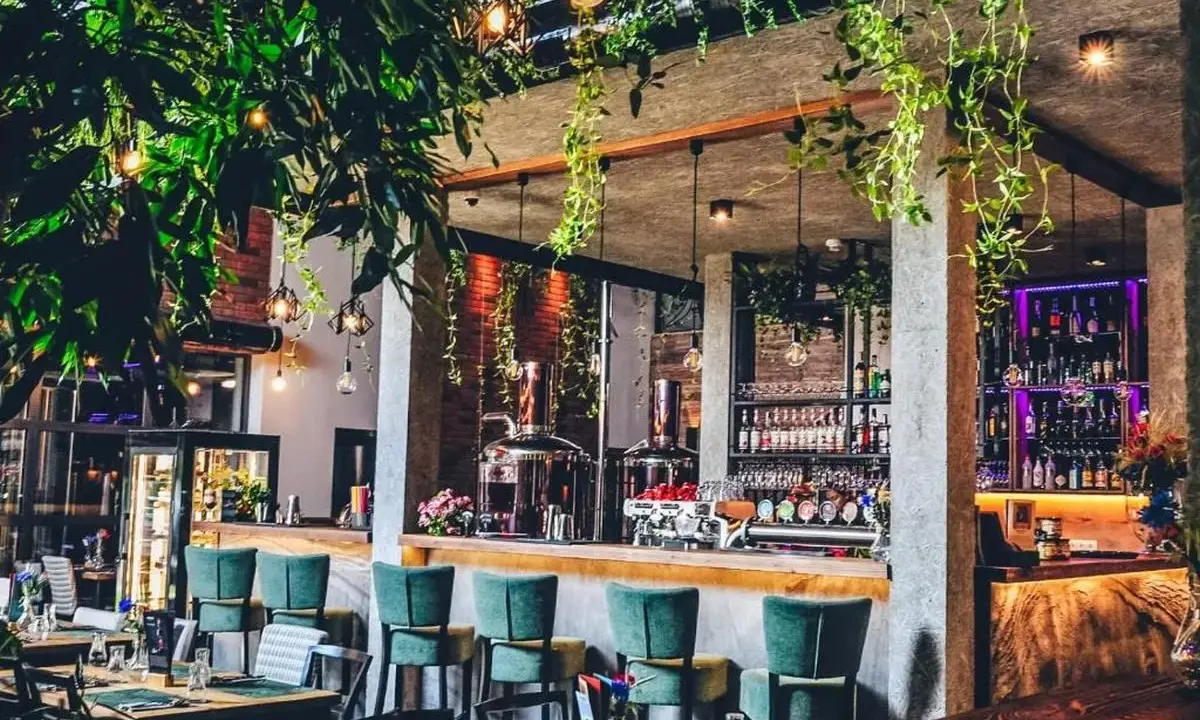 Wnętrze hotelu z browarem: bar z zielonymi hokerami, miedziany destylator, półki z butelkami i mnóstwo zieleni.