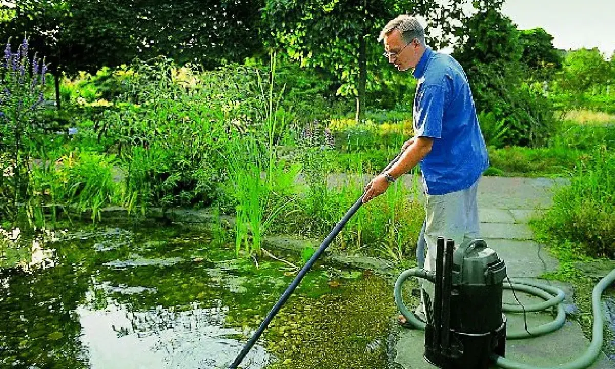 Mężczyzna wykonuje czyszczenie oczka wodnego za pomocą specjalnego odkurzacza. Wokół bujna zieleń.