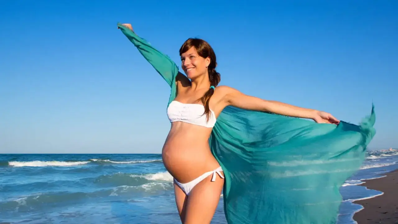 Donna incinta in bikini bianco sulla spiaggia, con un velo turchese che svolazza al vento. Il mare sullo sfondo.