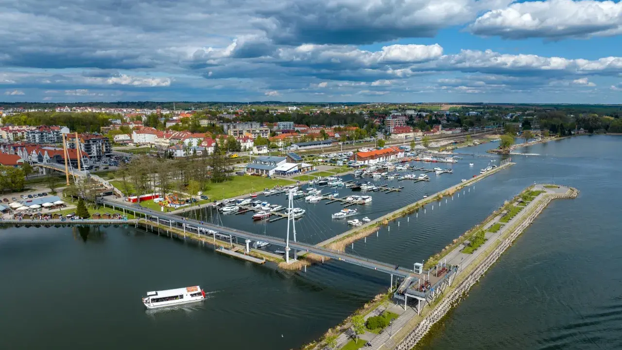 Widok na port w Giżycku, gdzie cumują liczne jachty i statki. Nowoczesny most pieszy łączy brzegi, a w tle rozciąga się miasto. To idealne miejsce na odkrywanie giżycko atrakcje.