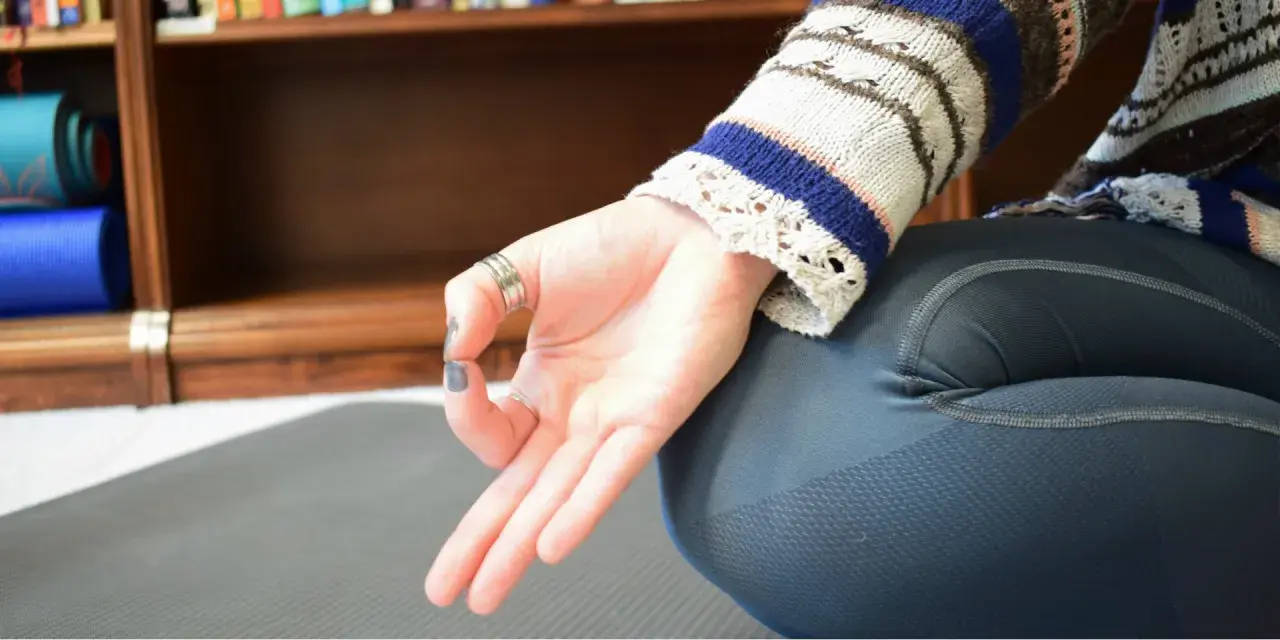 En hand utför en mudra, en gest som används i yoga och meditation. Bakom syns en bokhylla med yogamattor.