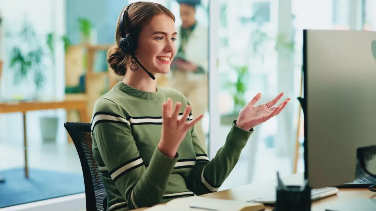 Uśmiechnięta customer experience manager w słuchawkach gestykuluje, siedząc przy biurku przed monitorem.