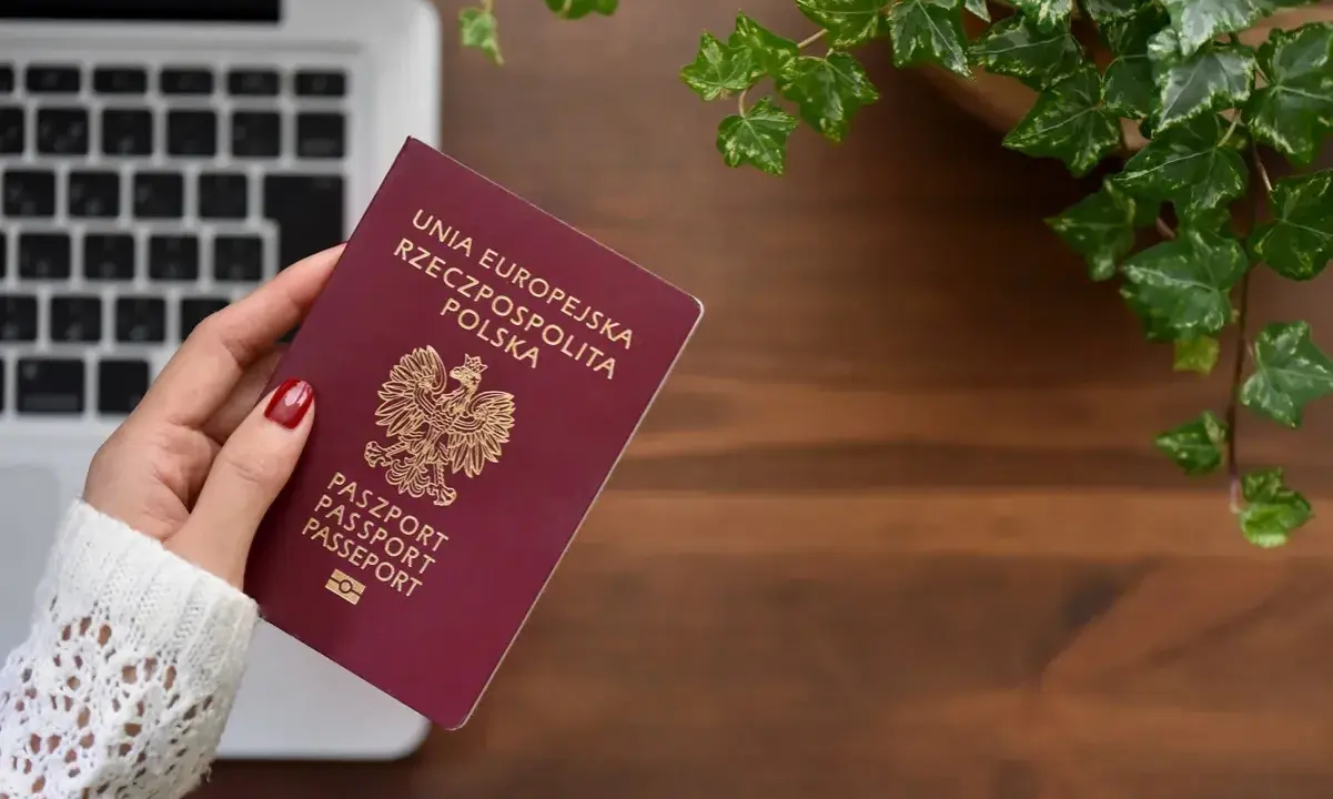Dłoń trzyma polski paszport na tle laptopa i roślin.