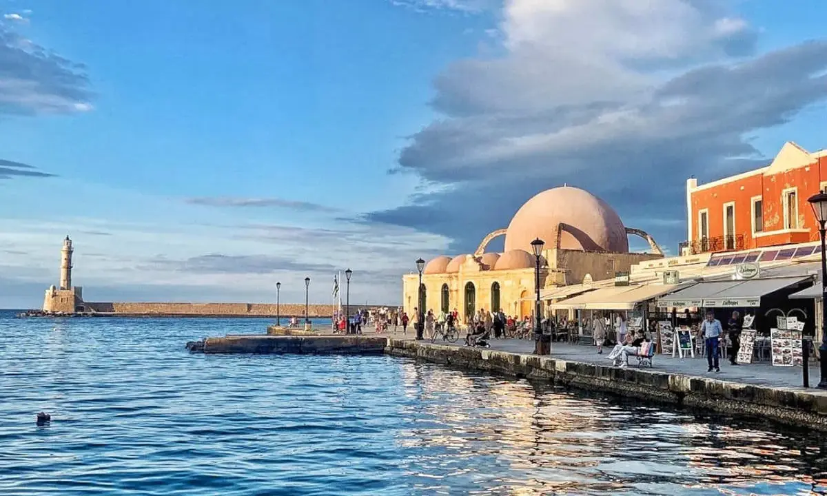 Il porto di Chania con il suo faro iconico e la moschea ottomana, bagnato da un mare blu intenso sotto un cielo nuvoloso.