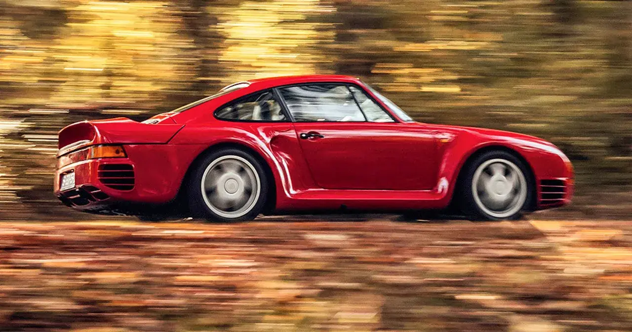 Czerwony Porsche 959 pędzi przez jesienny las, liście tworzą rozmyte smugi.