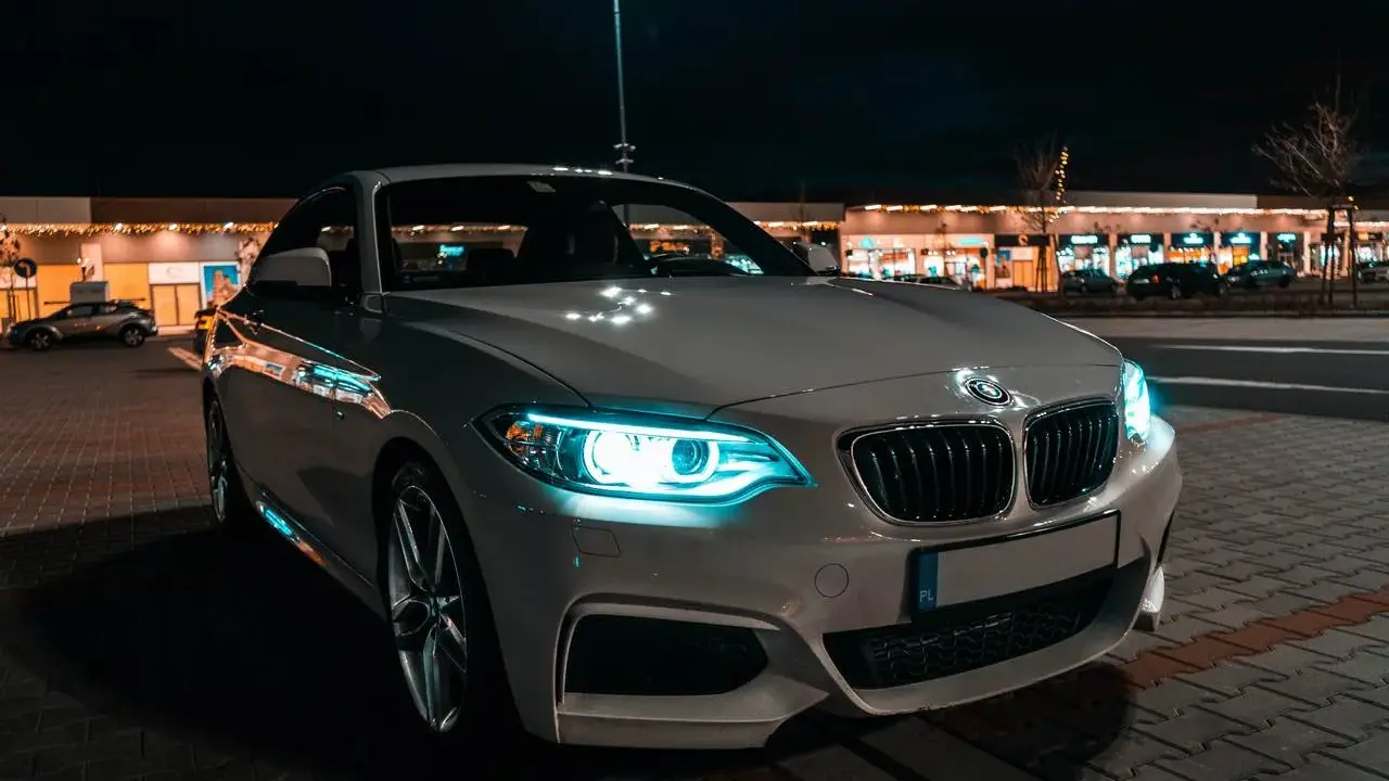 Białe BMW z włączonymi światłami adaptacyjnymi błyszczy nocą przed sklepami.