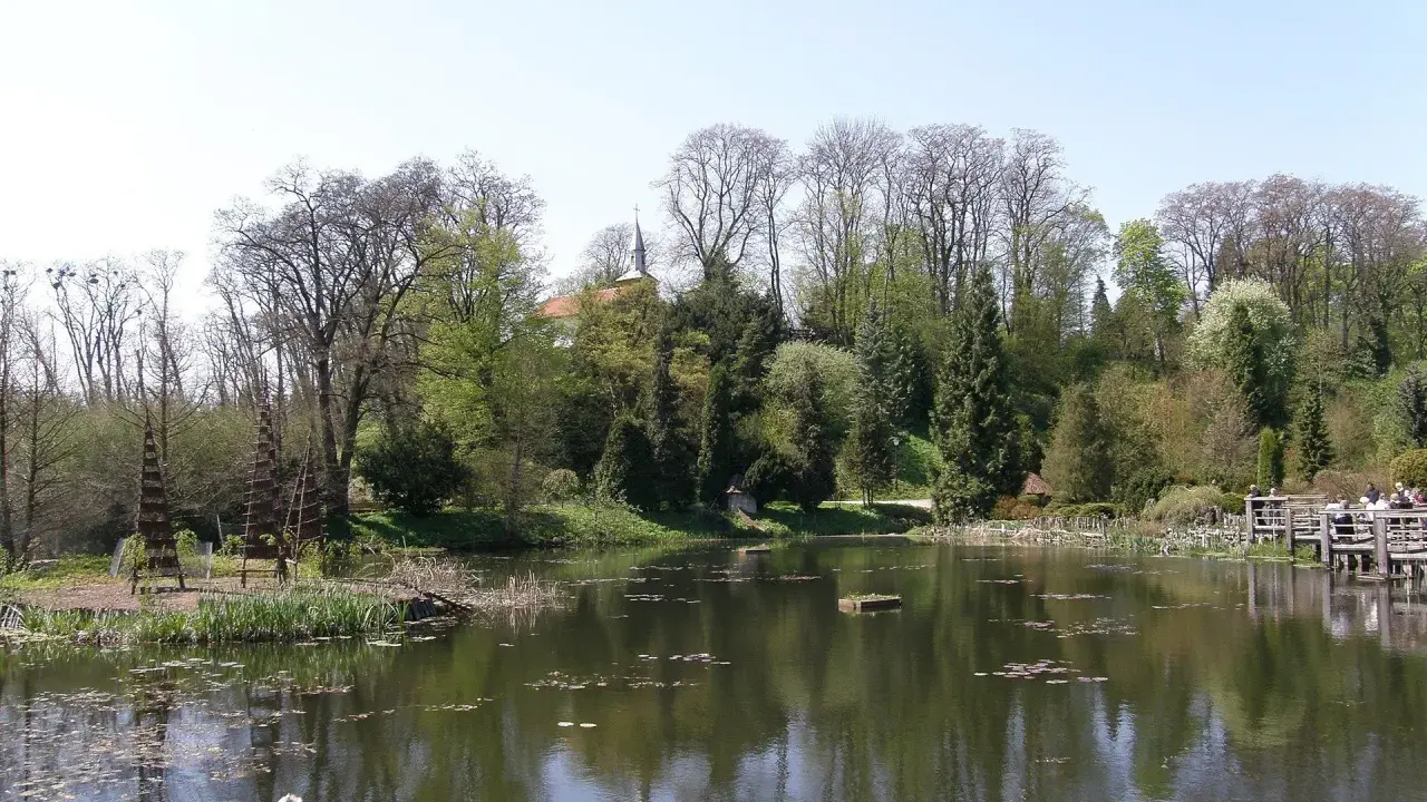 Spokojny staw w Bolestraszycach, otoczony drzewami i zielenią. Widoczna drewniana kładka z ludźmi, idealne miejsce na relaks i podziwianie atrakcji.