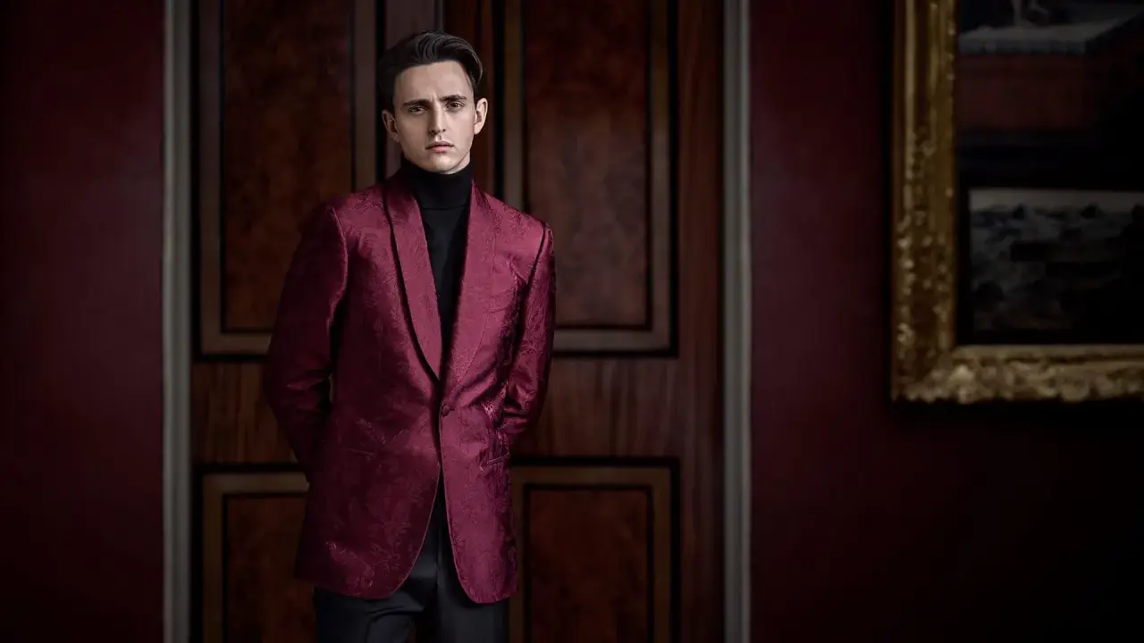 A man in a patterned burgundy jacket and black turtleneck, perfect for cocktail party attire for men, stands in a dimly lit room with ornate wooden doors.