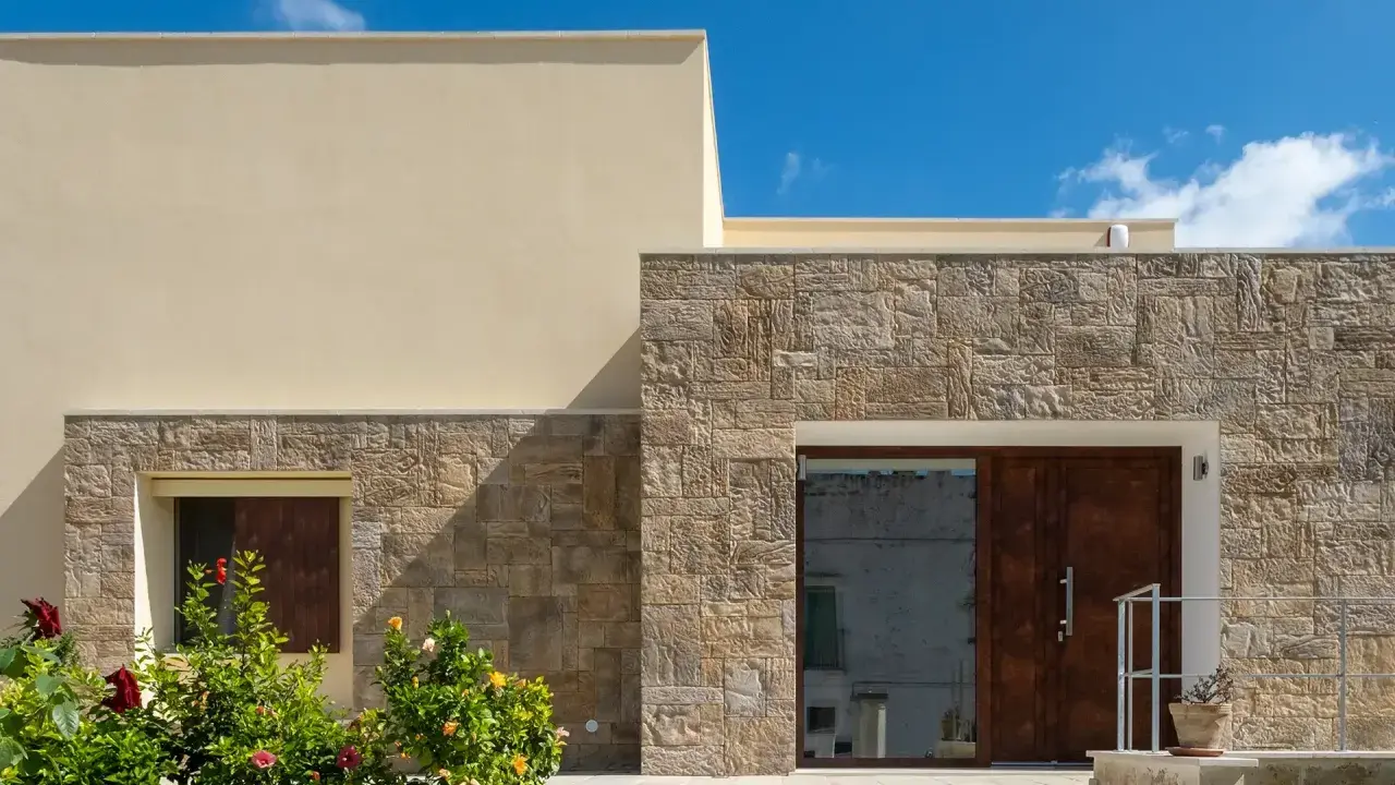 Moderne facciate in pietra grezza si alternano a intonaco chiaro, creando un contrasto materico. Un ingresso con porta in legno scuro e un giardino fiorito.