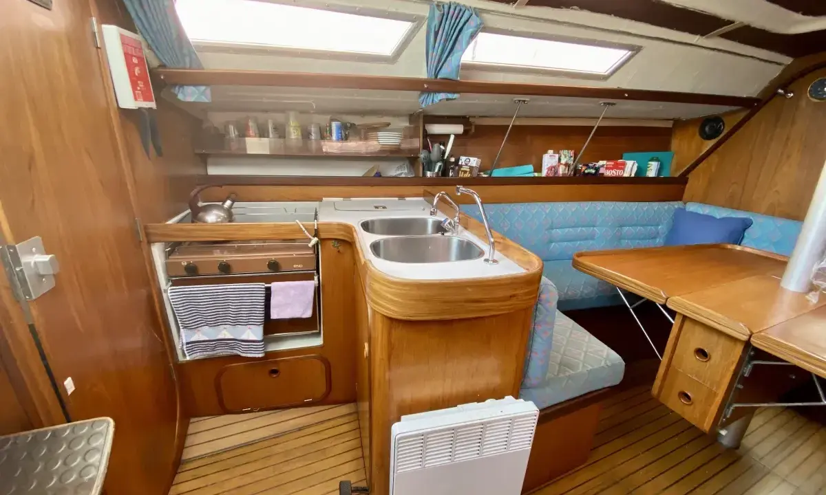Cuisine de voilier avec cuisinière, évier double, banquette bleue et table en bois. Un radiateur est visible. 312 avis gib sea.