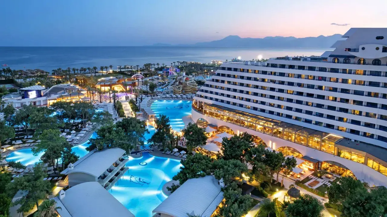 Luksusowy hotel Titanic Deluxe Lara z basenami i parkiem wodnym, oświetlony wieczorem, z widokiem na morze i góry.