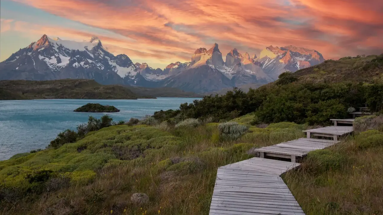 Drewniana ścieżka prowadzi przez zielone wzgórza ku turkusowemu jezioru i ośnieżonym górom. Zachód słońca maluje niebo nad **Tierra del Fuego** odcieniami różu i pomarańczy.