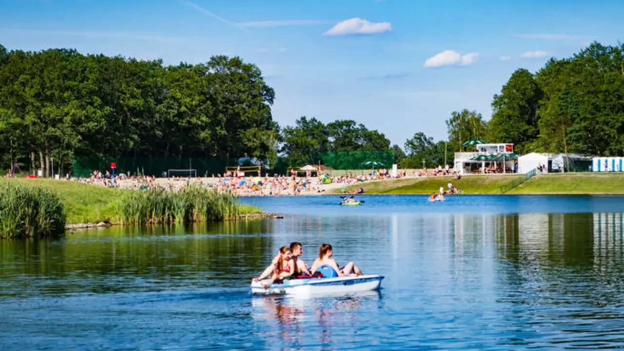 Rodzina pływa rowerkiem wodnym po jeziorze na Dolnym Śląsku, w tle plaża pełna ludzi i zielone drzewa.