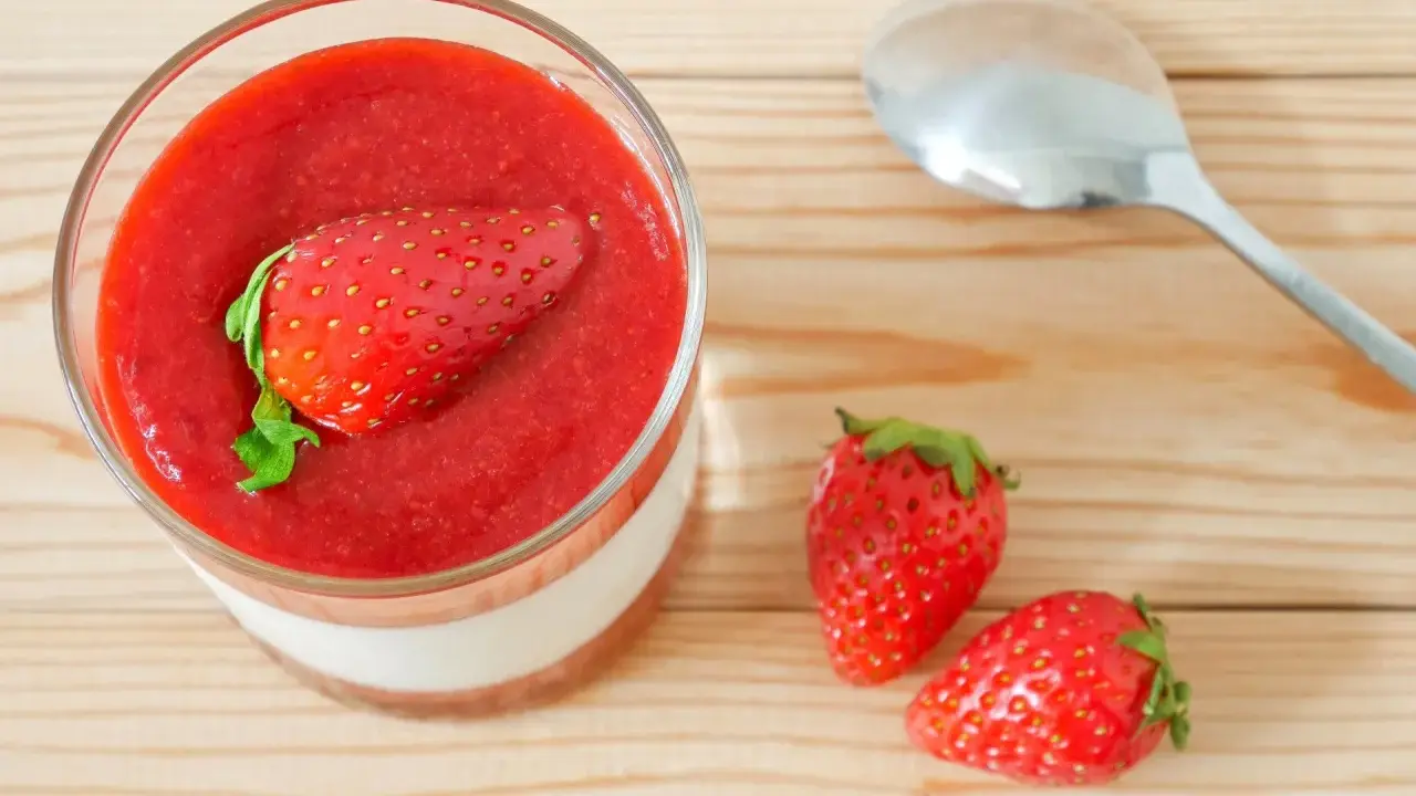 Un verre de panna cotta avec une fraise entière sur un coulis de fraise. Deux fraises fraîches et une cuillère à côté.