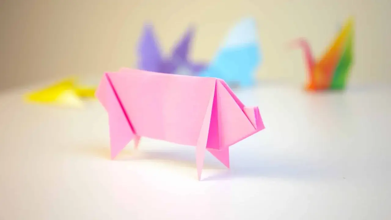 Cerdo de origami rosa con otras figuras de papiroflexia desenfocadas al fondo.