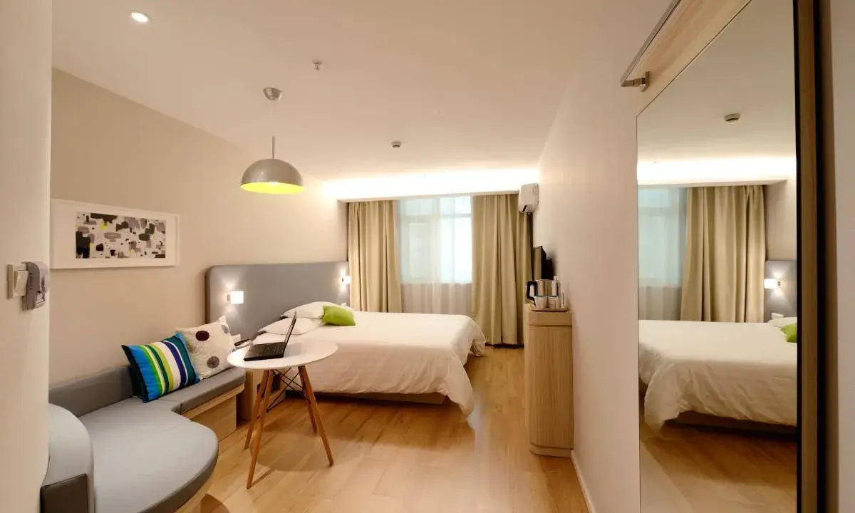 Habitación estrecha y alargada con cama, sofá curvo, mesa con portátil y espejo de cuerpo entero.