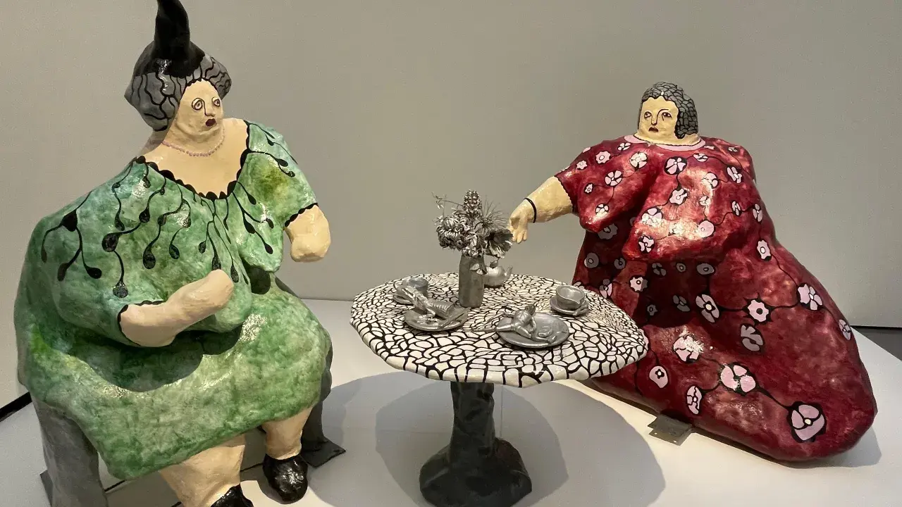 Dos figuras voluminosas, al estilo de Niki de Saint Phalle, sentadas a una mesa de té. Una viste de verde, la otra de rojo.