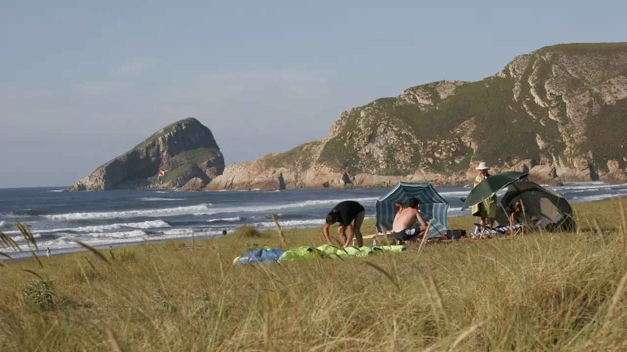 Gente en un playón de bayas, con carpas y sombrillas, disfrutando del mar y las montañas.