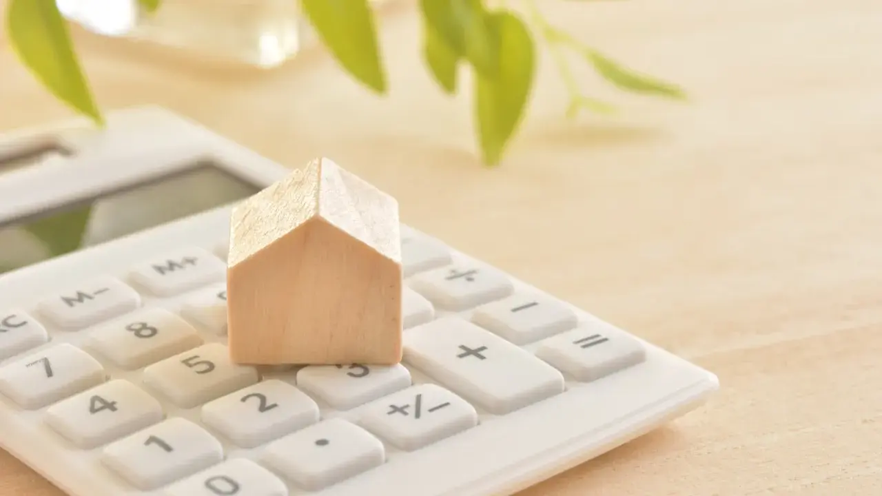 Maison en bois sur une calculatrice blanche, symbolisant le calcul du prix du **pel plafond**. Des feuilles vertes floues en arrière-plan.