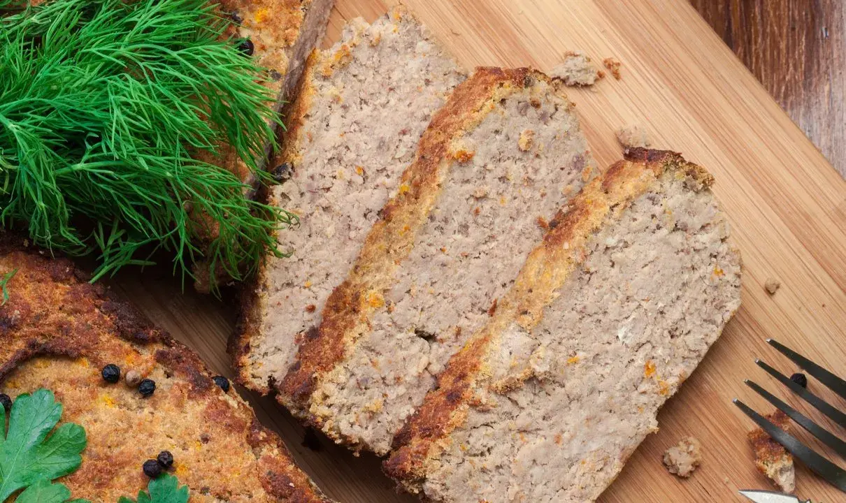 Pokrojony pasztet drobiowy z wątróbką, ozdobiony koperkiem i natką pietruszki, czeka na desce do krojenia.