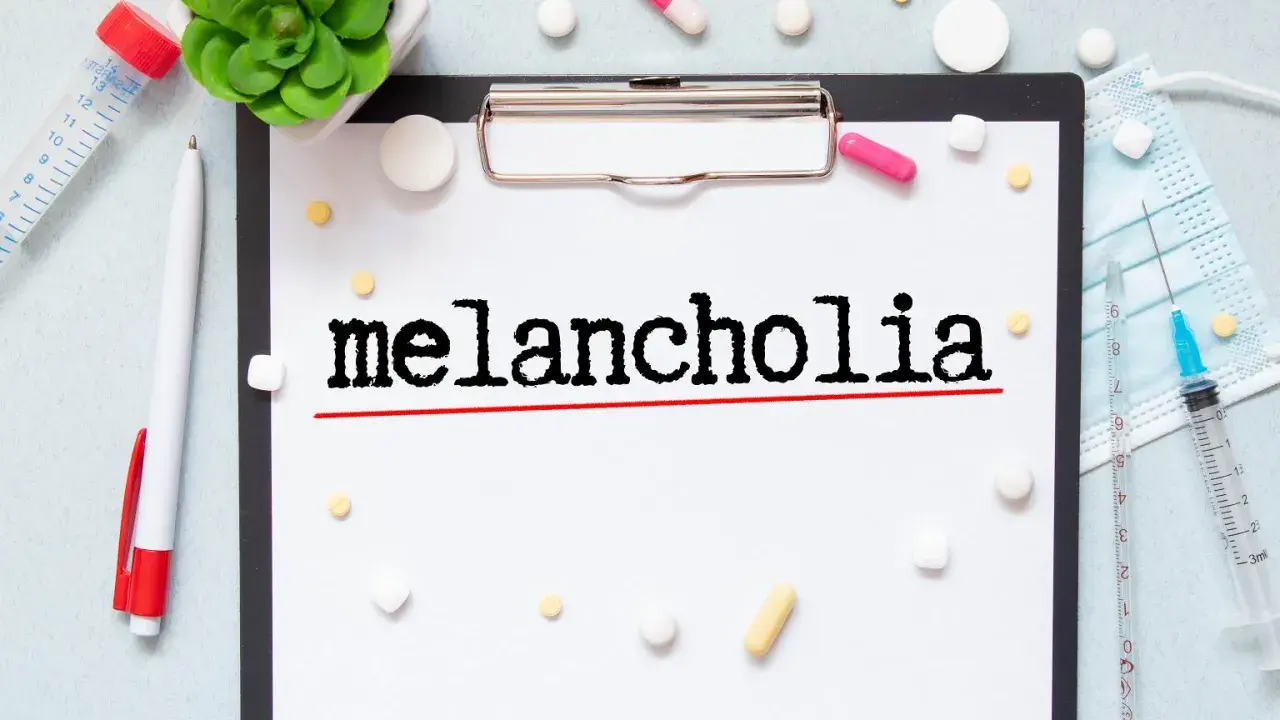 Na kartce widnieje napis "melancholia". Wokół leżą tabletki, strzykawka i maseczka, sugerując walkę z tym stanem.
