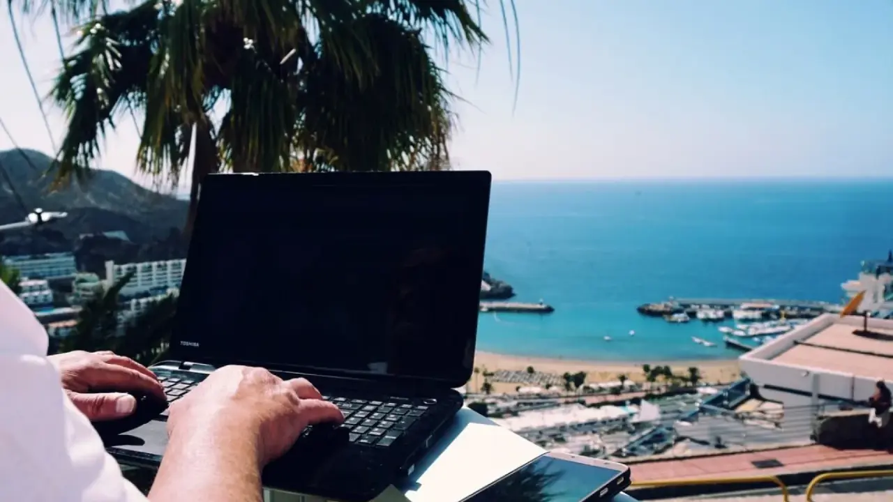 Praca na Cyprze: człowiek pisze na laptopie z widokiem na morze i palmę. Idealne miejsce na zdalną pracę.