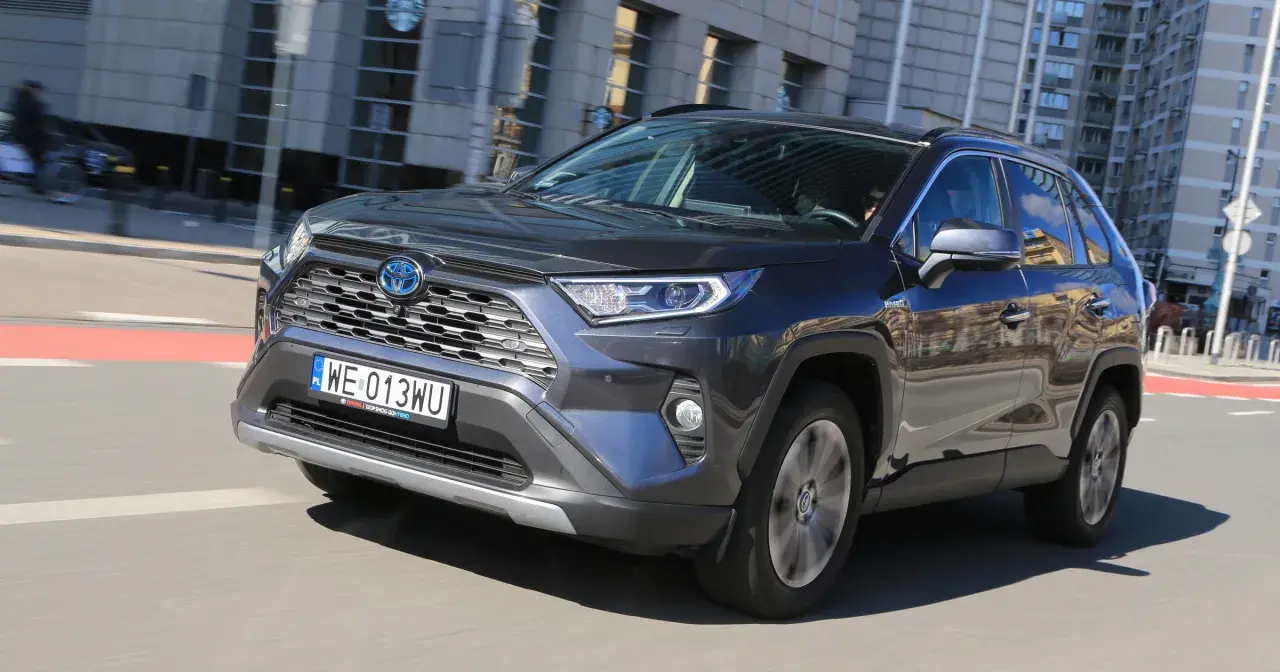 Ciemnoszary Toyota RAV4 Hybrid pędzi po mieście, odbijając w lustrach budynki i niebo.