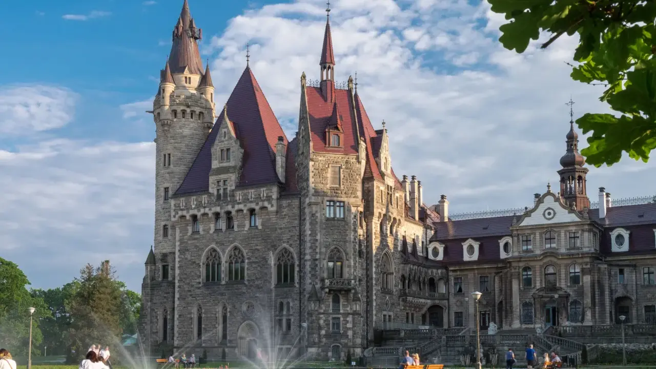 Imponujący zamek a pałac z kamienia, z wieżami i czerwonymi dachami, otoczony zielonym parkiem, gdzie odpoczywają ludzie.