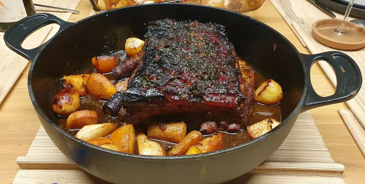 Poitrine de porc rôtie, croustillante et savoureuse, servie avec des pommes de terre et des carottes dans une cocotte en fonte.