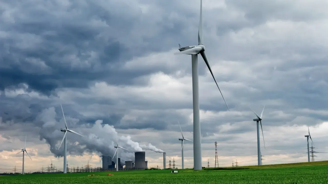 Windräder auf grüner Wiese unter dramatischem Himmel, im Hintergrund rauchende Industrieanlagen. Ein Symbol für Investitionen in erneuerbare Energien in Deutschland.