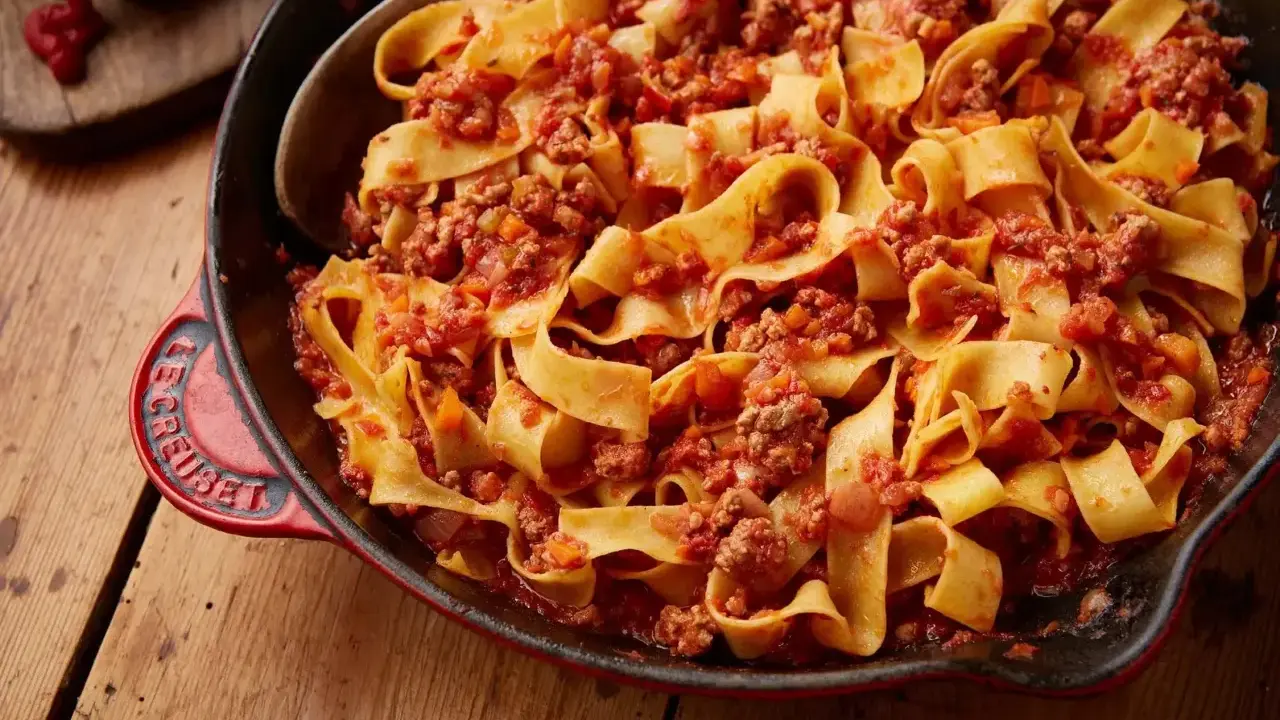 En stor portion pasta bolognese med breda bandnudlar och en mustig köttfärssås i en röd gjutjärnspanna.