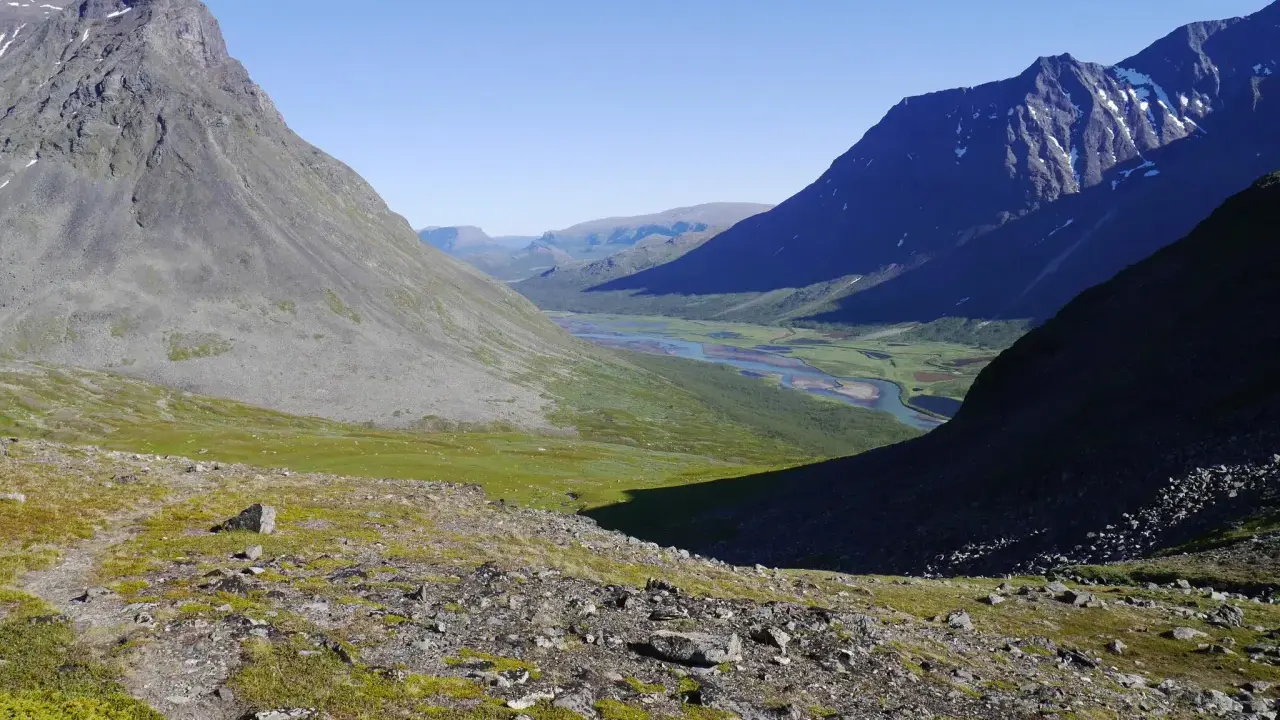 Vacker vy över Sareks nationalpark med Rapadalen i fjärran. En flod slingrar sig genom en grön dal omgiven av höga, steniga berg under en klarblå himmel.