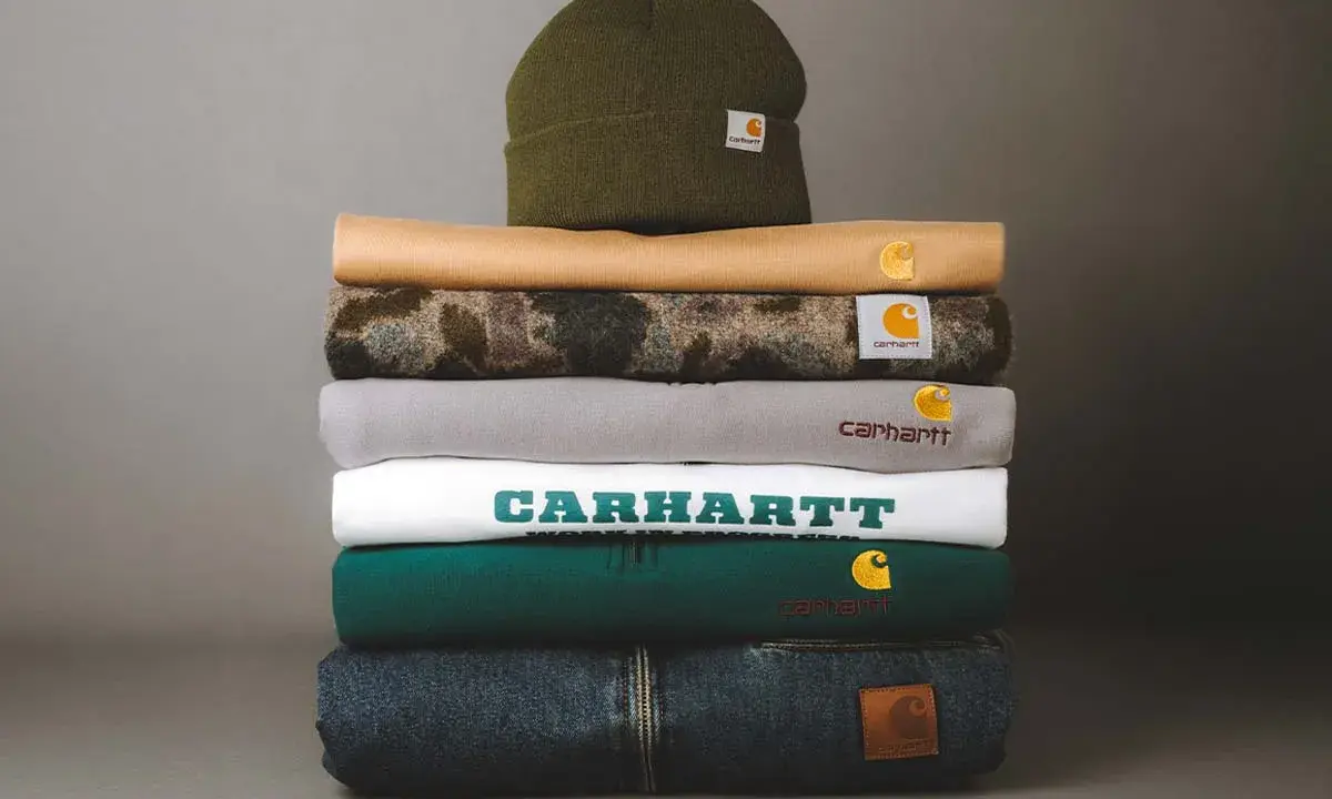 Stos ubrań Carhartt: czapka, bluzy i jeansy. Carhartt to firma znana z wytrzymałej odzieży roboczej i streetwearowej.
