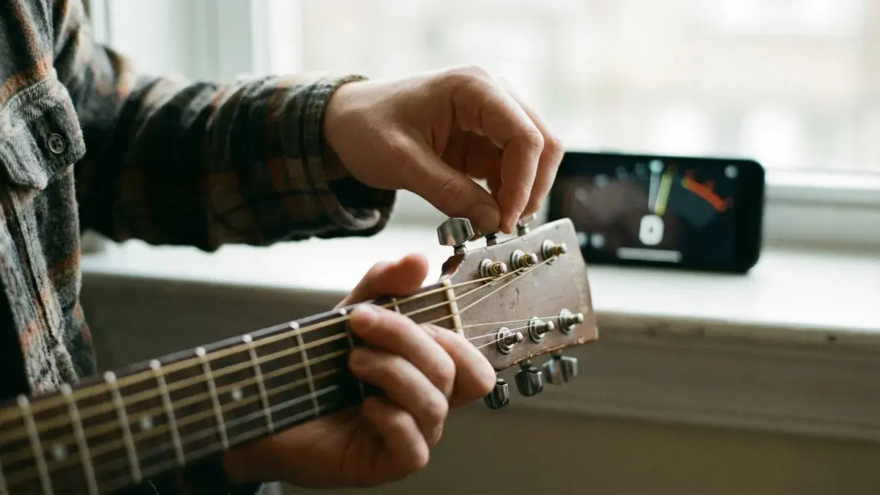 Main d'un musicien ajustant une guitare accordée, avec un accordeur sur un téléphone en arrière-plan.