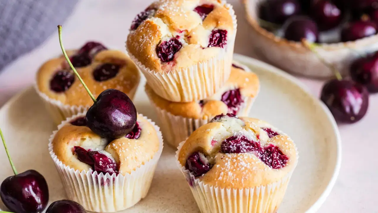 Saftige Muffins mit Sauerkirschen, bestäubt mit Puderzucker. Frische Kirschen liegen daneben und auf einem Muffin.