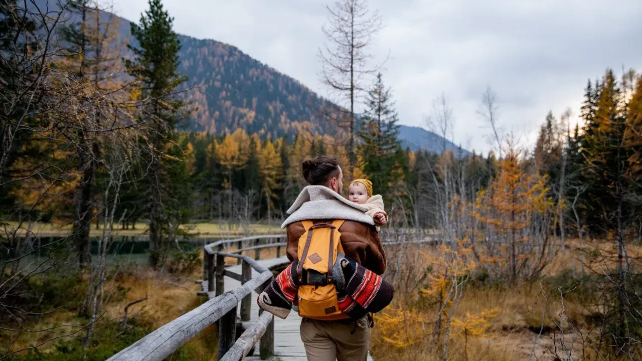 Ojciec z dzieckiem na plecach spaceruje drewnianą kładką pośród jesiennych lasów. Tatry z dzieckiem to piękna przygoda.