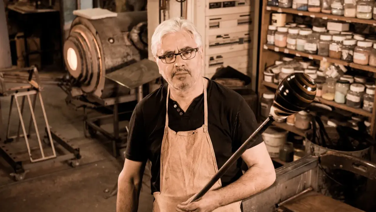 Un artisan verrier expérimenté, tablier en cuir, tient sa canne à souffler devant son four incandescent.