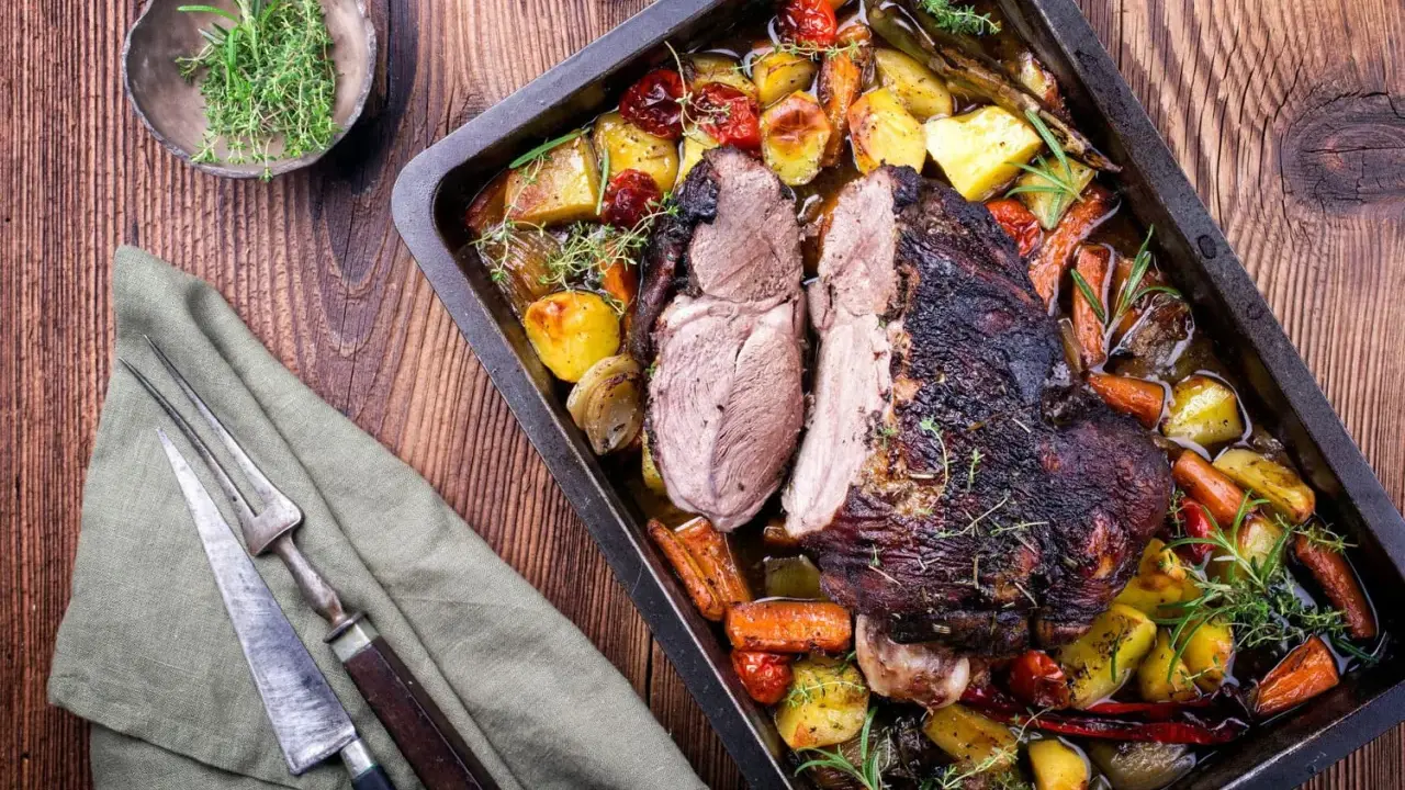 Gigot d'agneau rôti, coupé en deux, servi dans un plat avec des pommes de terre, des carottes et des herbes. Un plat savoureux prêt à être dégusté.