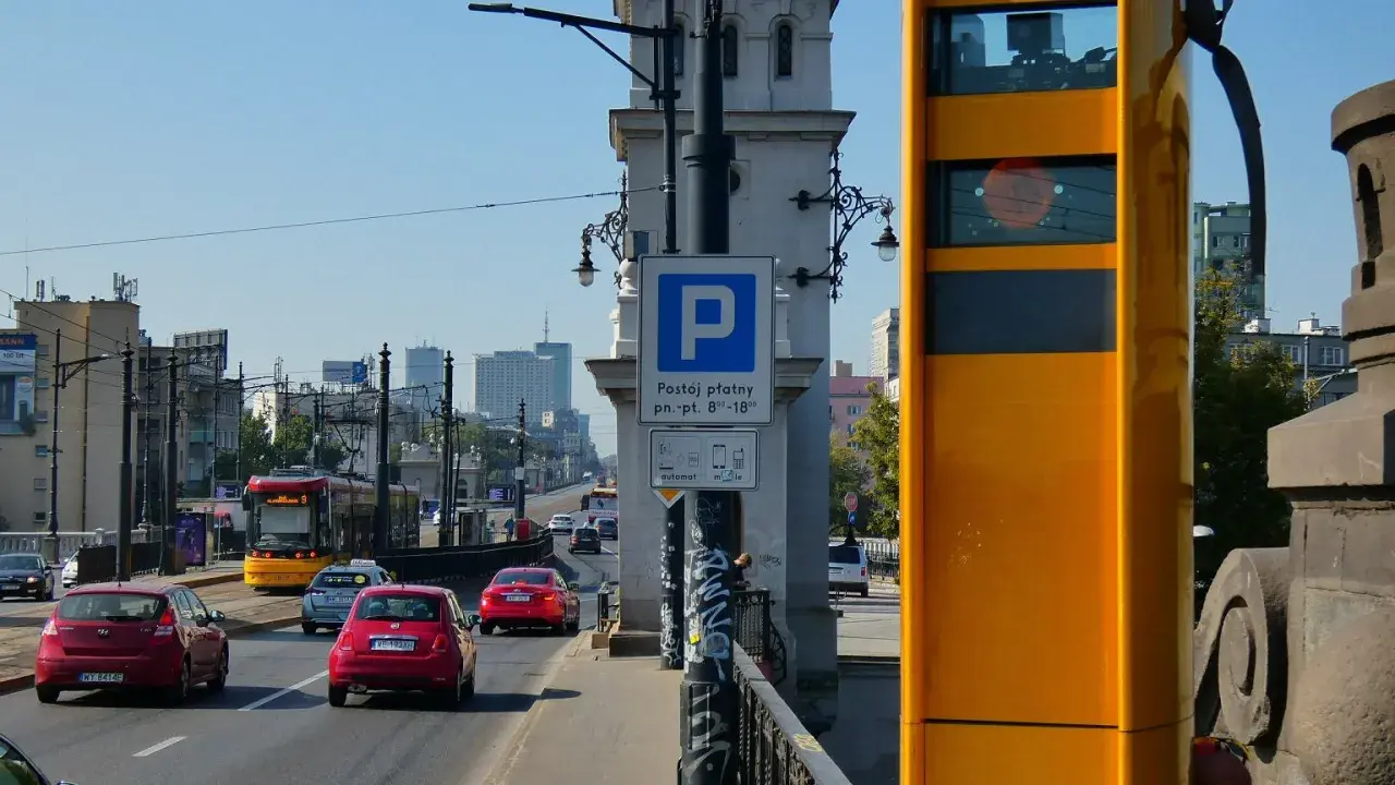 Żółty fotoradar stoi przy drodze, gdzie widać ruch samochodowy i tramwaj. Obok znak parkingu płatnego.