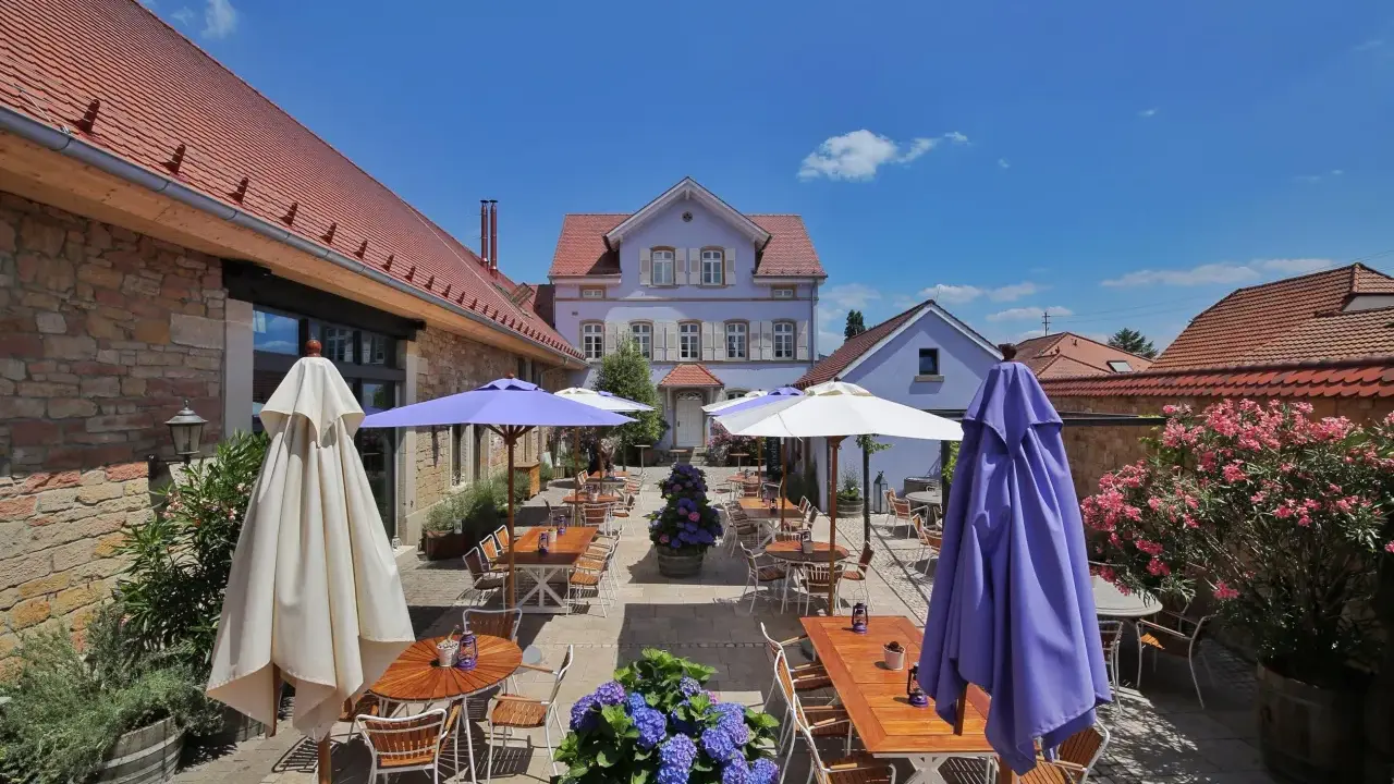 Terrasse eines Weinguts mit Tischen, Stühlen, Sonnenschirmen und Blumen unter blauem Himmel.