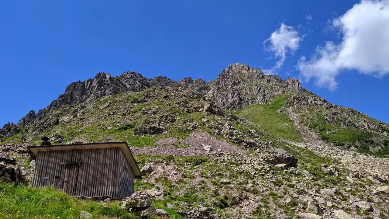 Bivacco Paolo e Nicola, un rifugio in legno ai piedi di montagne rocciose sotto un cielo azzurro.