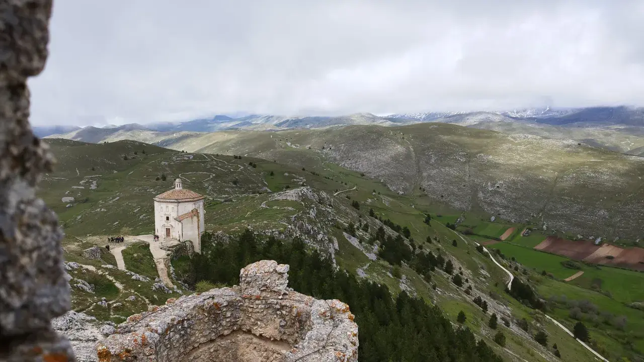 Un piccolo santuario domina il paesaggio del **cammino del Gran Sasso**, tra colline verdi e montagne nebbiose.