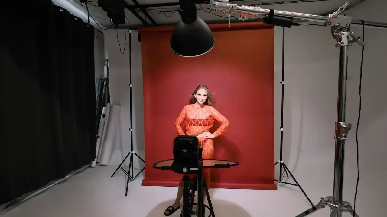 Modelka w pomarańczowej sukience pozuje na tle czerwonego tła. Wokół niej profesjonalne **światło studyjne** i sprzęt fotograficzny.